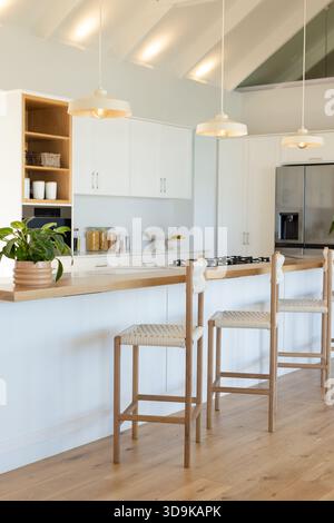 Helle Küche mit Insel-Arbeitsplatte mit gewebten Sitzhockern, Glasgläsern und Pendelleuchten Stockfoto