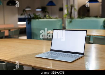Der schlanke silberne Laptop sitzt auf einem gemeinschaftlichen Holztisch in der Coworking Lounge mit blauen Kabinen Stockfoto