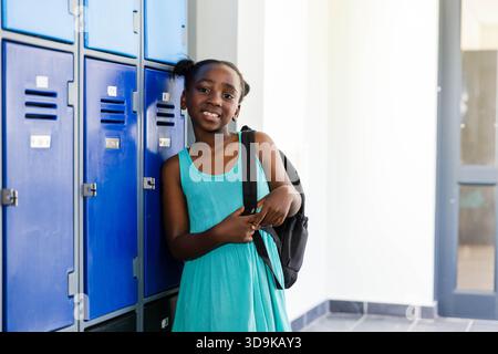 Weibliches Kind lehnt sich an blaue Schließfächer in der Schulhalle und hält Rucksack in türkisfarbenem Kleid Stockfoto