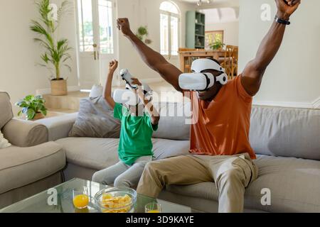 Afroamerikanischer Vater und Sohn mit weißen VR-Headsets, die den Controller auf dem Sofa im Wohnzimmer halten Stockfoto
