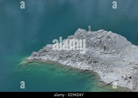 Eine kleine Schotterinsel erhebt sich aus hellem türkisfarbenem Wasser, umgeben von einer ruhigen Seenlandschaft. Die klare Oberfläche spiegelt die ruhige Umgebung wider, enha Stockfoto