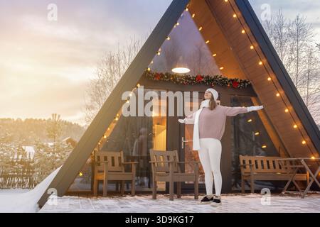 Frau genießt den Winteruntergang, während sie vor einer gemütlichen dreieckigen Hütte steht, die mit Lichtern in einer verschneiten Landschaft dekoriert ist Stockfoto