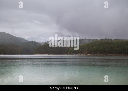 Eine ruhige Berglandschaft mit Nebel über den Wald und einem ruhigen See, der den bewölkten Himmel reflektiert. Stockfoto