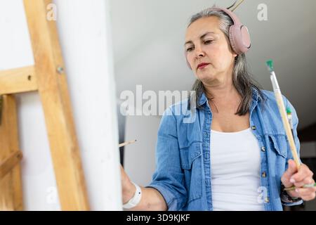 Malerin mit Pinseln, die Leinwand malen, während sie im Kunststudio pinkfarbene Kopfhörer trägt Stockfoto