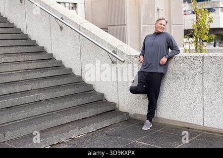 Senior Mann lehnt sich an die Betonwand an der plaza und blickt nach rechts, während er Smartwatch trägt, Kopierraum Stockfoto
