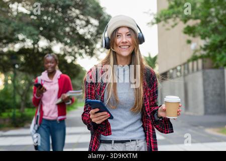 Verschiedene Studentinnen gehen auf dem Campus-Gehweg mit Kaffeetasse, Smartphones und Notebooks Stockfoto