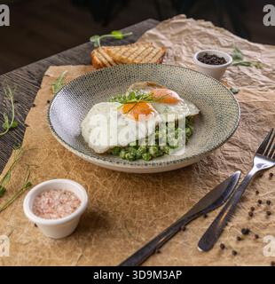Teller mit Spiegeleiern auf grünem Erbsenpüree, Toast und Gewürzen auf rustikalem Papier Stockfoto