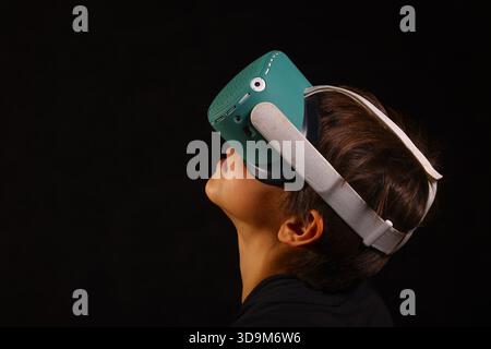 Junge, der ein VR-Headset trägt und nach oben blickt, konzentriert sich auf virtuelle Inhalte in dunkler Studioumgebung. Stockfoto