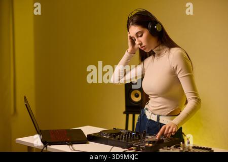 Weibliche DJ-Mischpulte auf Plattenspielern, farbenfrohe Lichtstudio, fokussierter Ausdruck, Hände einstellender Mixer und Laptop-Bedienelemente, Kopfhörer tragen, modern Stockfoto