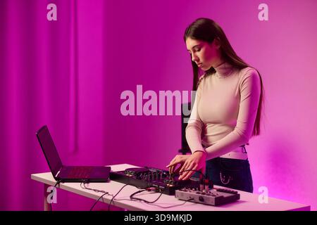 Weibliche DJ-Mischpulte auf Plattenspielern, farbenfrohe Lichtstudio, fokussierter Ausdruck, Hände einstellender Mixer und Laptop-Bedienelemente, Kopfhörer tragen, modern Stockfoto