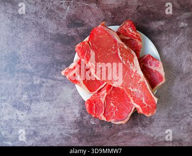 Blick von oben auf drei New York Strip Steaks auf einer weißen Platte auf einer Tischplatte mit Tageslicht. Stockfoto