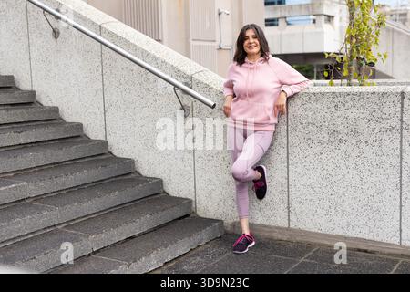Frau, die sich an eine niedrige Wand neben dem Treppenhaus auf dem stadtplatz lehnt und pastellfarbene Aktivkleidung trägt, Kopierraum Stockfoto