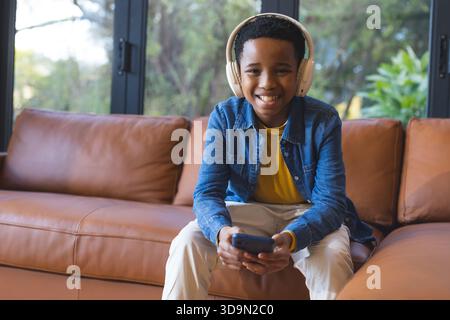 Afroamerikanischer Junge lächelt, während er das Smartphone im Wohnzimmer mit weißen Kopfhörern auf dem Sofa hält Stockfoto