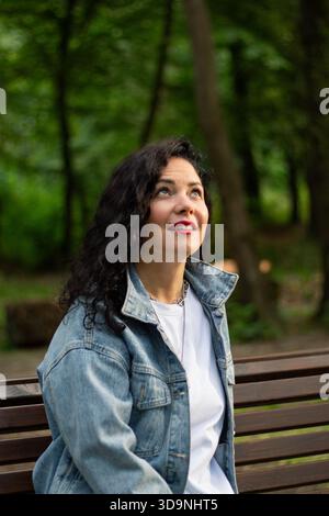 Porträt einer selbstbewussten lockigen Frau in der Natur. Nahaufnahme-Porträt einer dunkelhaarigen Frau mit voluminösen Locken, die selbstbewusst vor verschwommenem Grün lächelt Stockfoto