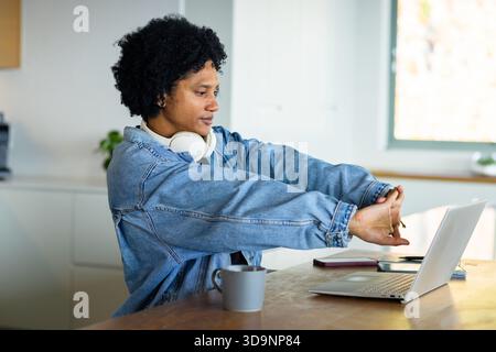 Mittelgroßer afroamerikanischer Mann, der die Arme nach vorne streckt, indem er in der Küche einen Laptop und Kopfhörer benutzt Stockfoto