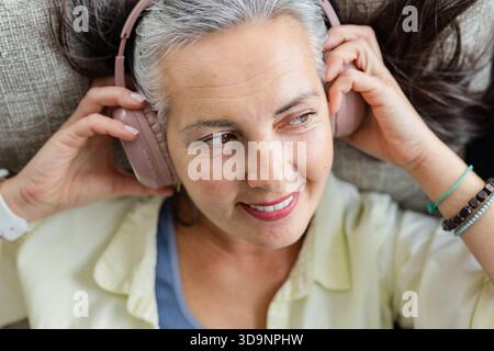 Frau, die sich auf einem grauen Sofakissen zurücklehnt und eine weiße Uhr trägt, die rosa kabellose Kopfhörer hält Stockfoto