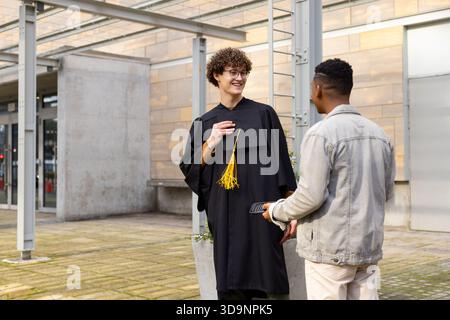 Verschiedene männliche Freunde im Kleid zeigen auf die Quaste und benutzen das Smartphone neben Pflanzgefäßen auf dem Campus Quad Stockfoto