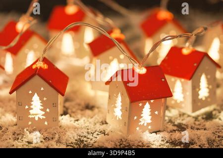Eine Schnur aus leuchtenden Miniaturlichtern. Weiße Weihnachtshäuser mit rotem Dach auf künstlichem Schnee Hintergrund, warme und gemütliche Winterferienszene Stockfoto