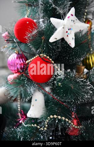 Aus nächster Nähe sehen Sie einen künstlichen Weihnachtsbaum, der hell mit flauschigen roten Kugeln, weißen Sternen und Gold- und Perlengirlanden verziert ist. Stockfoto