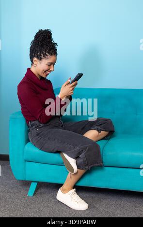Afroamerikanerin, die ein schwarzes Smartphone hält, während sie lächelnd auf einem türkisfarbenen Sofa in der Lounge sitzt Stockfoto