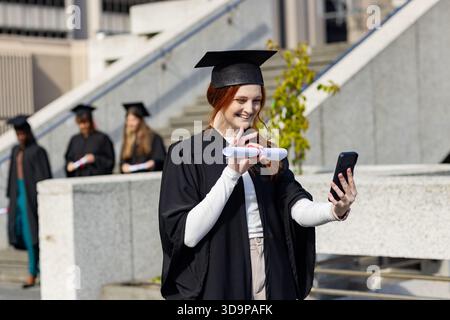 Verschiedene weibliche Klassenkameraden, die Kleider tragen, stehen auf dem Campus plaza, haben Diplome und winken am Telefon Stockfoto