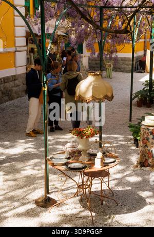 Triest, Italien - 20. April 2025: Holztisch in einem Gartenhof mit Vintage-Tellern, Krügen und Kerzenständern. Ein Gartenstuhl aus Metall und V Stockfoto