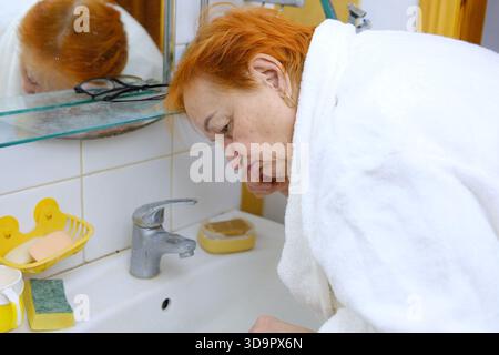 Rothaarige Frau in einem Gewand ältere Frau putzt sich vor dem Spiegel im Badezimmer die Zähne. Zahnhygiene, Vitalität und Schönheitskonzepte. Hochwertige Fotos Stockfoto