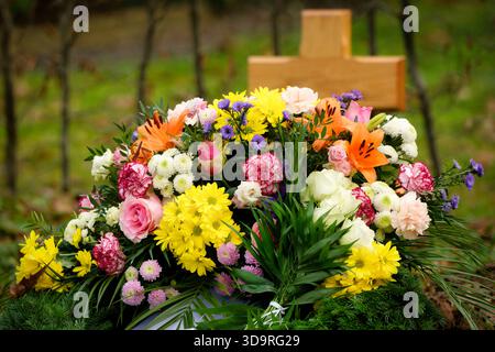 Farbenfrohe, üppige Blumenarrangements mit hellen Lilien, gelben und weißen Chrysanthemen und rosa Rosen, die auf einem Grab mit einem Holzkreuz ruhen Stockfoto