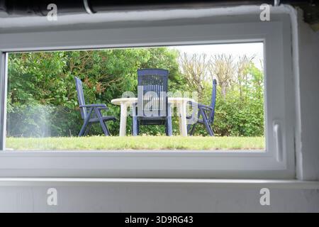 Drei schwarze Plastikstühle und ein kleiner Tisch sitzen draußen auf Gras, umgeben von grünen Büschen, durch ein Plastikfenster gesehen Stockfoto