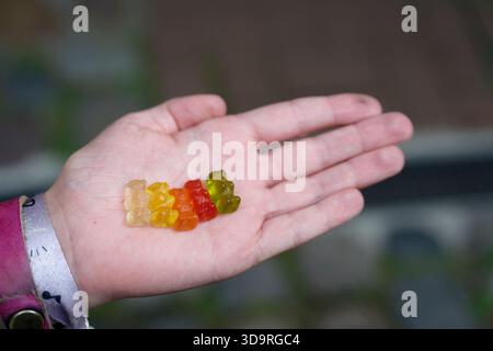 Eine Kinderhand hält fünf Gummibärchen in grün, rot, orange, gelb und klar. Es werden zwei Armbänder getragen – ein Band, ein rosafarbenes Leder mit Nieten. Stockfoto