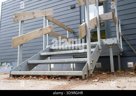 Metalltreppe mit vier Stufen, die zu einer schwarzen Glastür führen, Holzschienen, die auf Metallstangen befestigt sind, trockene Blätter und Schmutz, der unten in einer Außenumgebung sichtbar ist Stockfoto