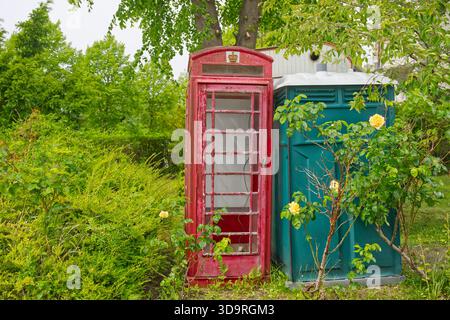 Eine alte rote britische Telefonzelle neben einer modernen grünen tragbaren Toilette, umgeben von üppigem Grün und gelben Blumen. Stockfoto