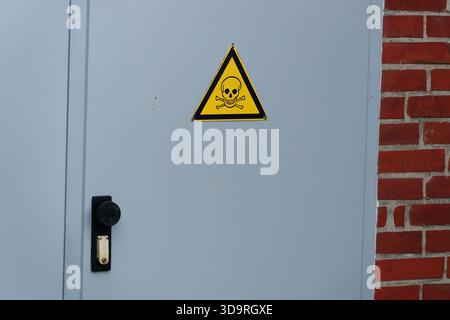 Eine Metalltür auf einem roten Backsteingebäude zeigt ein lebhaftes gelbes dreieckiges Schild mit einem Schädel und gekreuzten Knochen, um toxische oder lebensbedrohliche Gefahren anzuzeigen Stockfoto