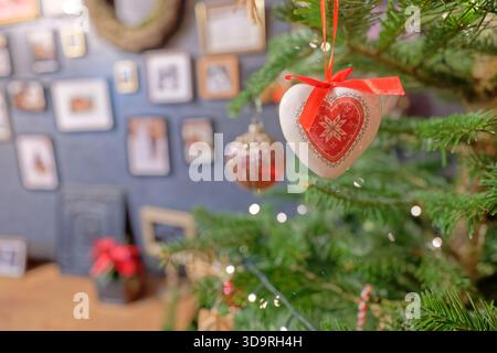 Ein weißes herzförmiges Ornament mit einem gemalten Herzen hängt an einem roten Band an einem verzierten Weihnachtsbaum, mit Bilderrahmen im gemütlichen Hintergrund. Stockfoto