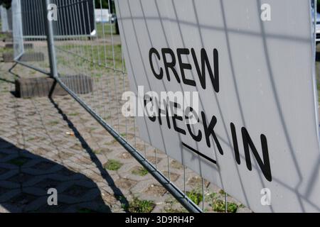 Ein Metallzaun zeigt ein weißes Schild mit der Aufschrift „Crew Check in“ in schwarzen Buchstaben. Grünes Gras wächst an einem Sommertag zwischen Fußbodenfliesen. Stockfoto
