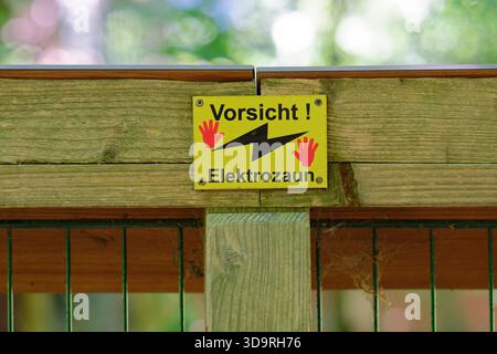 Ein gelbes Warnschild mit der Aufschrift Vorsicht Elektrozaun ist auf einer Holzplatte über einer Metallbarriere montiert, die Teil eines umzäunten Tiergeheges mit ist Stockfoto