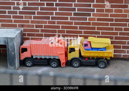 Zwei Spielzeugwagen aus Kunststoff, ein roter und ein gelber mit Kippbetten, befinden sich auf dem Boden in der Nähe einer roten Ziegelwand und einer Betonbank mit einem kleineren Mauerwerk Stockfoto