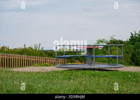 Ein Metallspielplatz steht auf einem grasbewachsenen Feld mit blauen Sitzen und einem roten Lenkrad. Im Hintergrund sind Holzzäune, Wildblumen und ein Stockfoto