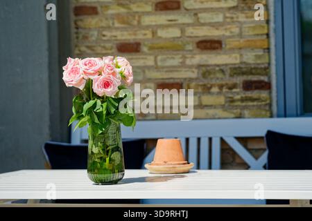 Auf einem weißen Tisch befindet sich eine Vase aus klarem Glas, gefüllt mit frischen rosa Rosen und grünen Blättern. Neben der Vase ist ein Aschenbecher. Die Einstellung befindet sich vor einem bei Stockfoto