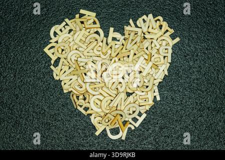 Eine Gruppe kleiner englischer Holzbuchstaben ist sorgfältig in Herzform auf einem schwarzen texturierten Teppich angeordnet, umgeben von anderen verstreuten Buchstaben für Stockfoto