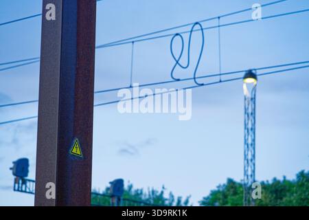 Ein rostiger alter Metallmast zeigt ein gelbes dreieckiges Warnschild mit einem Blitz, der Elektrizität anzeigt. Im Hintergrund, Straßenlaternen und rai Stockfoto
