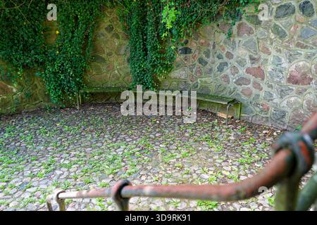Eine hohe Steinmauer aus großen Steinen in Beton verläuft entlang des Weges drei alte Bänke, deren Oberflächen mit Moos vergilbt sind Stockfoto