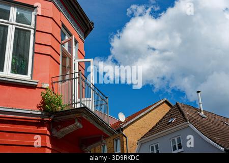 Die obere Etage ist hellrot und die untere Fassade in hellem Rot. Sie zeigen einen charmanten alten Balkon mit Metallgeländer und offenen weißen Holztüren. Häuser in der Nähe Stockfoto