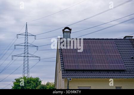 Ein hellgelbes Haus mit einem dunklen Fliesendach, ausgestattet mit Solarpaneelen und einem Kamin steht neben einem Strommast mit gestreckten Drähten, die eine Mischung aus Mod schaffen Stockfoto