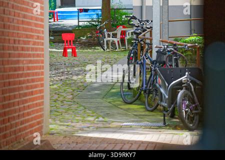 Ein schmaler, von Fahrrädern gesäumter Steinweg umfasst einen mit einem Kinderanhänger, während in der Mitte ein hellroter Holzstuhl steht, umgeben von weißem pla Stockfoto
