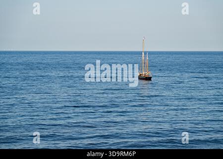 Ein kleines Segelboot mit Segeln nach unten gleitet friedlich über ein blaues, ruhiges Meer, während der Himmel darüber rein und klar erscheint, ohne Wolken, die eine Ruhe schaffen Stockfoto