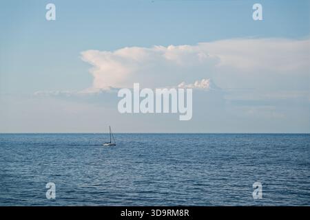 Eine kleine Yacht mit Mast und abgesenktem Segel bewegt sich leise über ein ruhiges blaues Meer, während einige weiße Wolken den klaren Himmel darüber schmücken und einen Friedenspaß schaffen Stockfoto