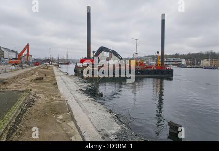 An der Flensburger Uferpromenade wird eine neue Promenade gebaut. Ein Arbeitsschiff trägt einen Bagger in Küstennähe, während Bewehrungsstahl aus frischem Beton herausragt Stockfoto
