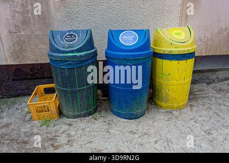 Auf einem Betonhof stehen drei hölzerne Mülltonnen, die gelb, blau und dunkelblau gefärbt sind. Auf den Etiketten sind Bio- und Restmuell-, Papier- und Gelbersack-Etiketten zu lesen. Gelb Stockfoto