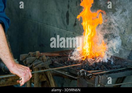 Ein offenes Kohlebett atmet eine heftige orangene Flamme. Ein Arbeiter erhitzt einen Stahlstab im Herzen des Feuers, dreht ihn für eine gleichmäßige Farbe und bereitet den Stack vor Stockfoto